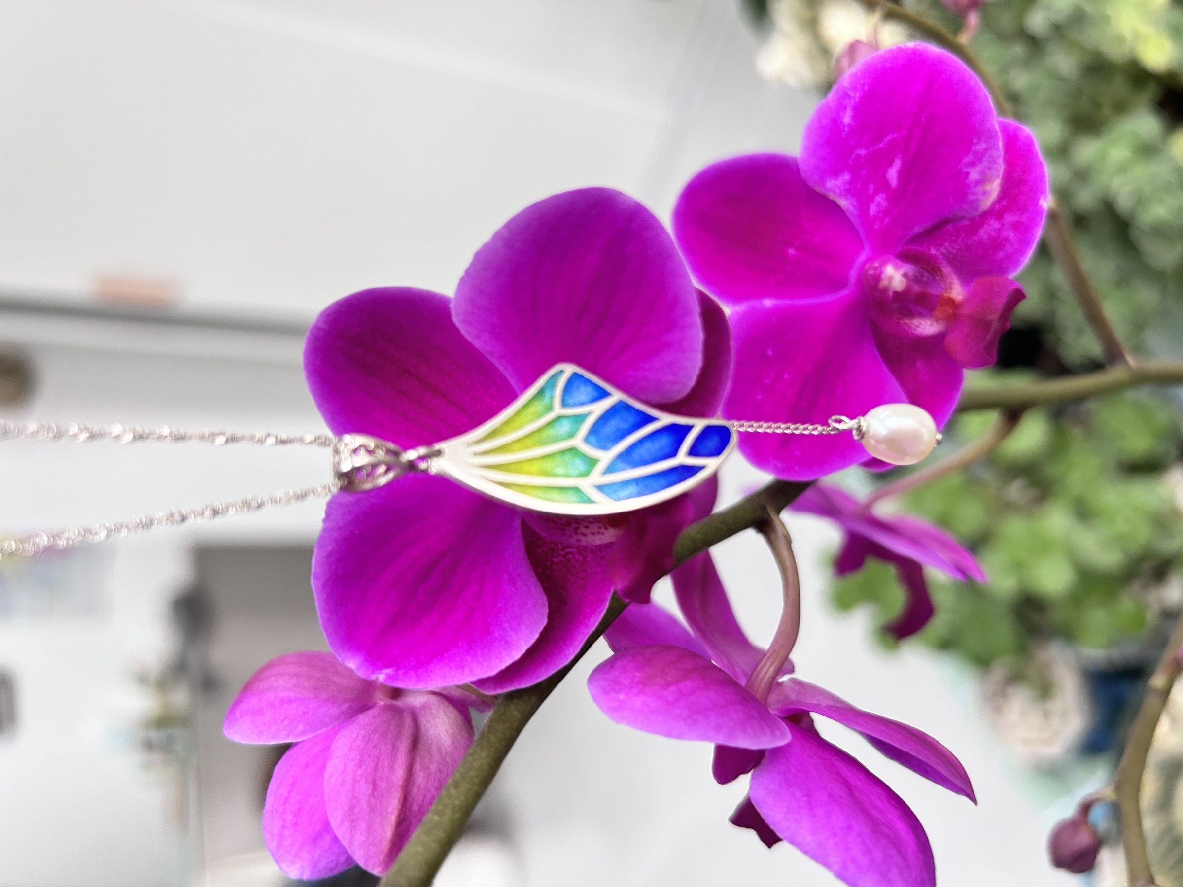 A plique-à-jour enamel butterfly-wing pendant resting on orchid petals — YÀN Atelier