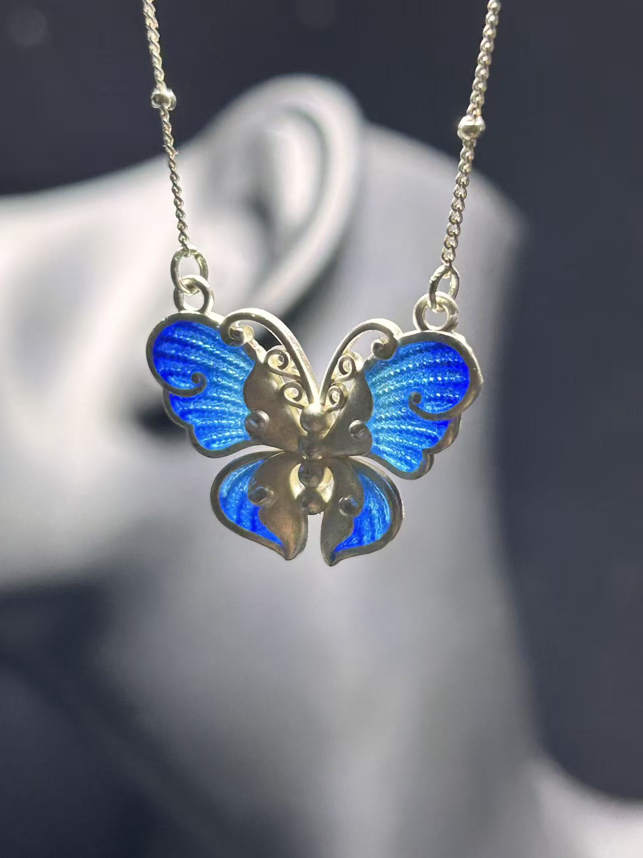 A blue plique-à-jour butterfly held to the light — translucent enamel, made at the atelier